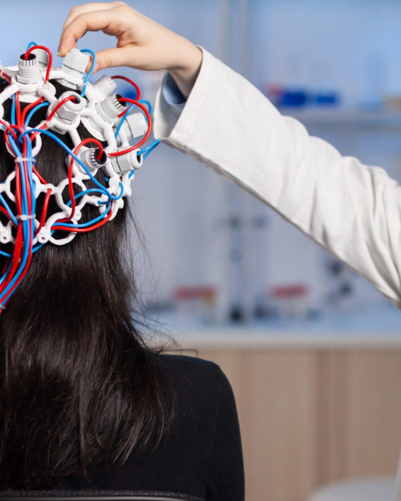 Back view of woman patient wearing performant eeg headset sitting on chair in neurological research laboratory while medical researcher adjusting it, examining nervous system typing on tablet.